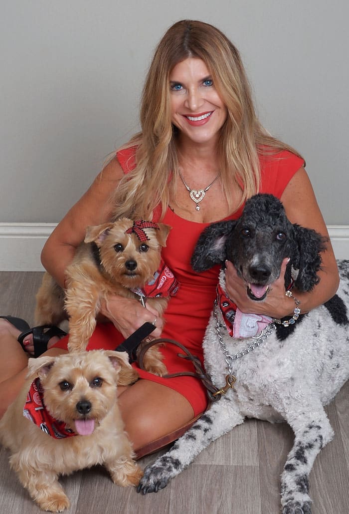 A smiling woman in a red dress sits on the floor with three dogs. Two small tan dogs and a larger black-and-white dog wear matching red bandanas. The group appears happy and relaxed against a plain light gray wall.