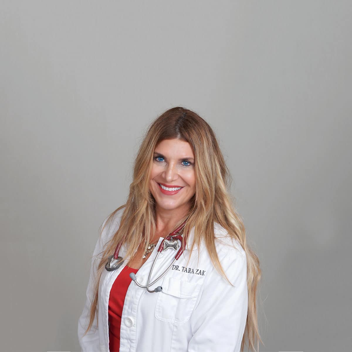 A smiling woman with long blonde hair, wearing a white lab coat with "Dr. Tara Zak" embroidered on it and a stethoscope around her neck, stands in front of a plain light gray background.